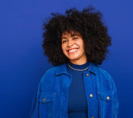 Feeling good in blue. Happy woman with curly hair smiling at the camera while standing against a blue background. Beautiful woman with curly hair wearing her natural air with pride.